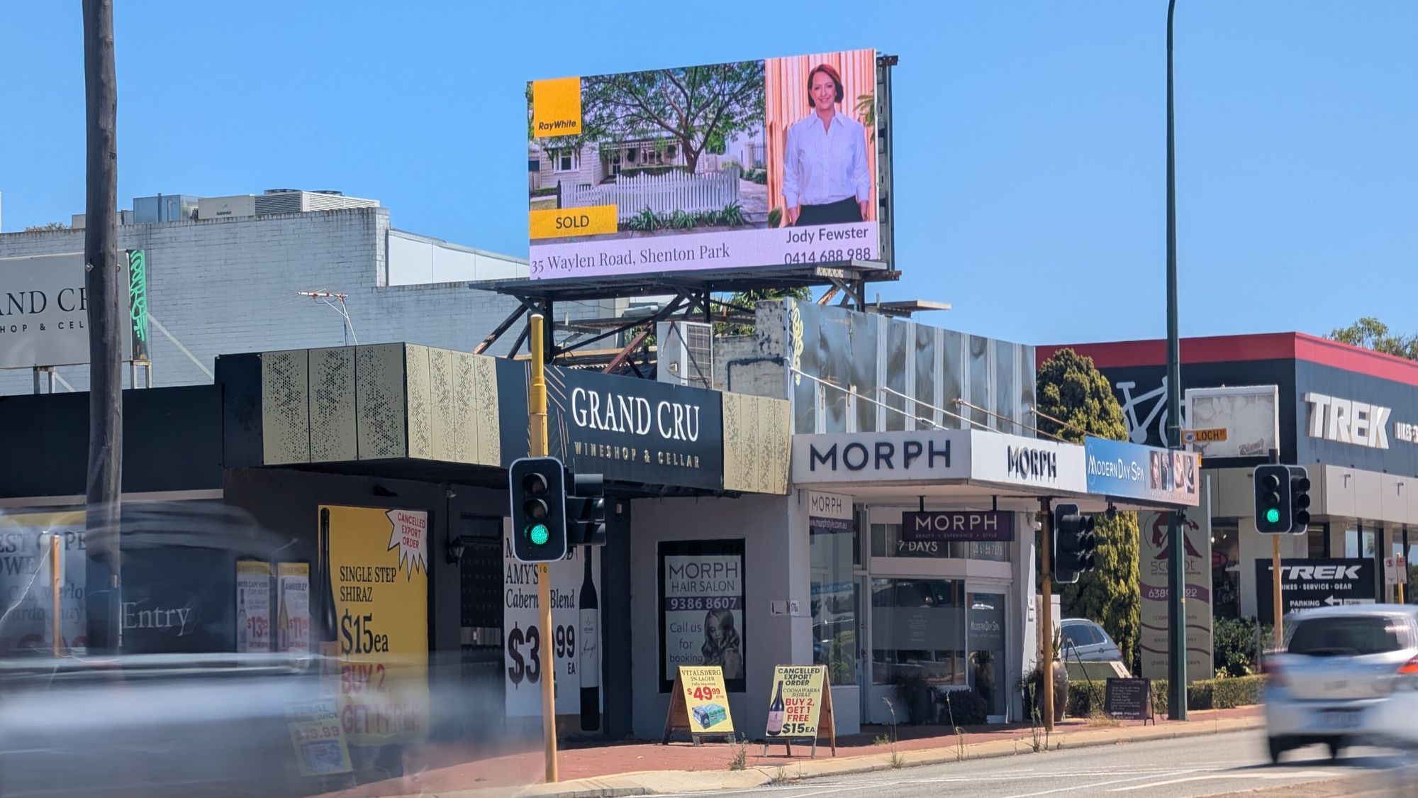 A digital billboard above shopfronts on Stirling Highway, Nedlands, with traffic passing.
